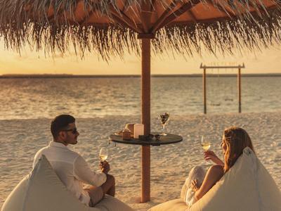 Drei Personen sitzen bei Sonnenuntergang auf Sitzsäcken unter einem Strohhutschirm am Strand.
