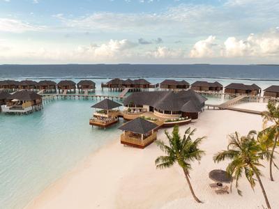 Luxuriöse Überwasserbungalows und Palmen an einem weißen Sandstrand mit klarem türkisfarbenem Wasser.