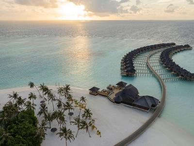 Bungalows auf Stelzen über klarem Wasser bei Sonnenuntergang mit Palmen und Sandstrand.