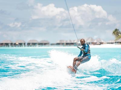 Mann wakeboardet auf klarem blauem Wasser mit Bungalows im Hintergrund.