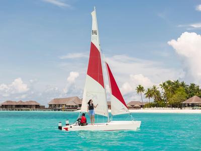 Segelboot mit roten und weißen Segeln auf türkisblauem Meer vor einer Insel mit Palmen.