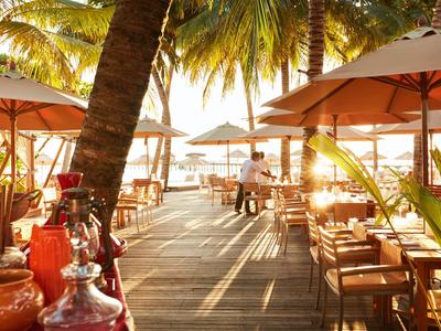Sonnige Terrasse eines Strandrestaurants mit Holzboden, Tischen, Stühlen und Palmen.