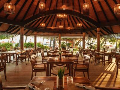 Offenes Restaurant mit Holztischen, Stühlen und dekorativen Lichtern unter einem Holzdach.