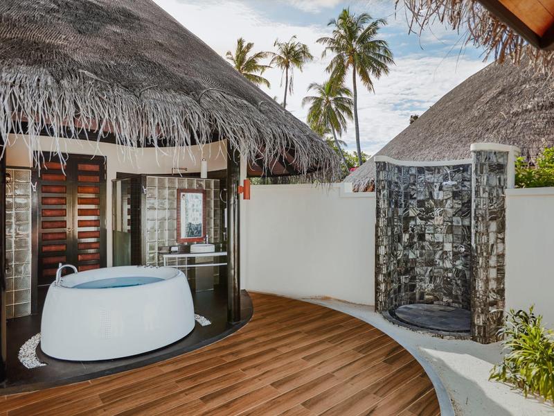 Zona de bienestar al aire libre con jacuzzi y ducha bajo techos de palma sobre terraza de madera.