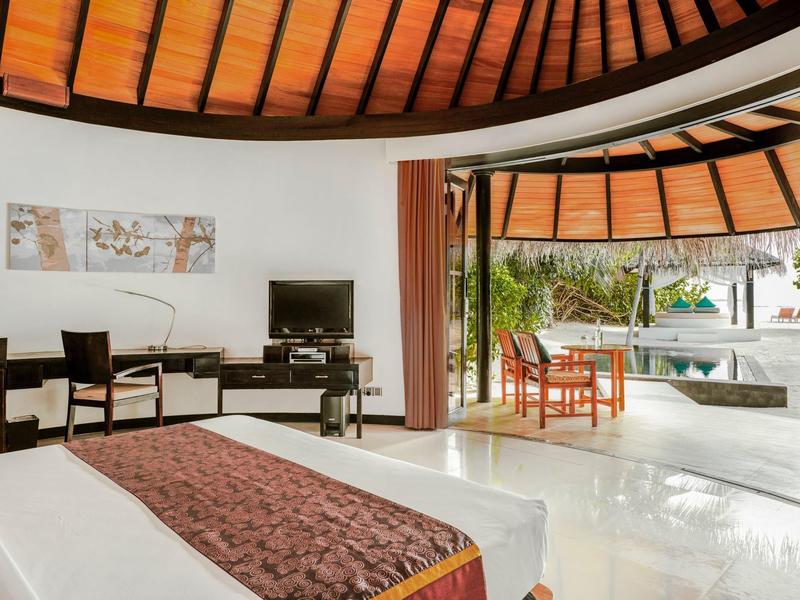 Habitación de hotel luminosa con techo de madera, cama, zona de estar y terraza con vista a la piscina.