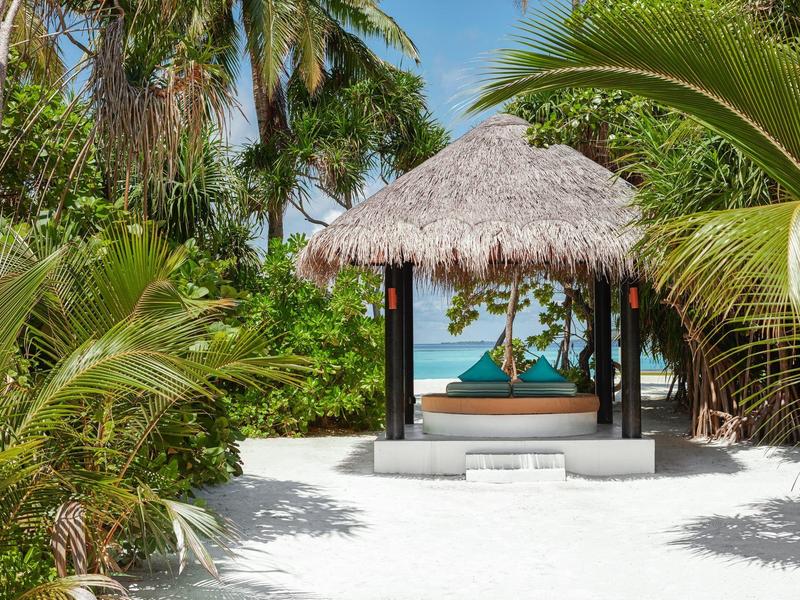 Un pabellón de techo de paja en playa de arena blanca con vista al mar turquesa, rodeado de palmeras.
