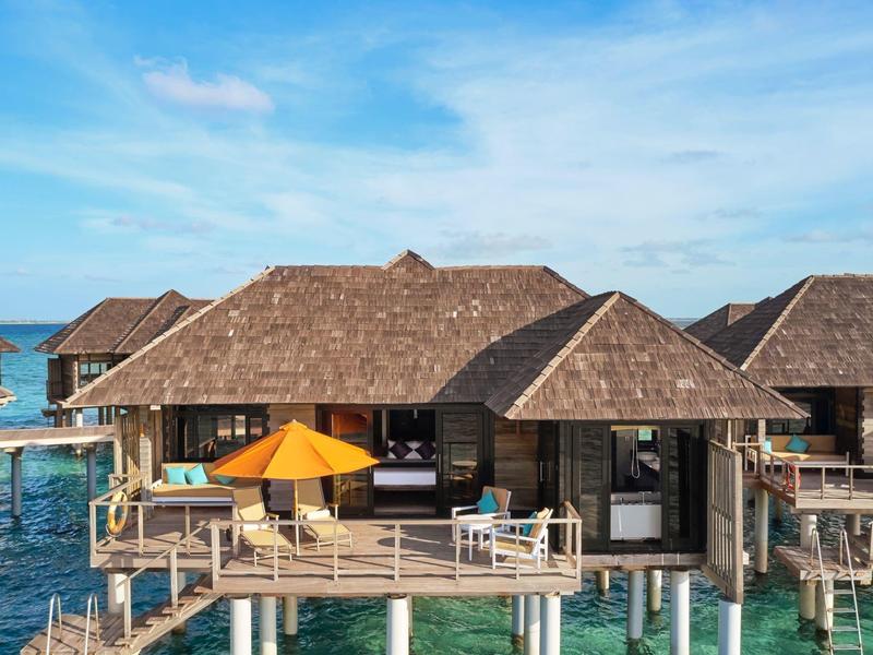 Bungaló sobre el agua con terraza, sombrilla naranja y vista al mar azul bajo un cielo despejado.