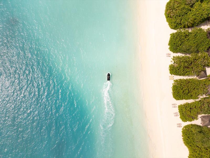 Vista aérea de un bote navegando en agua turquesa clara junto a una playa de arena blanca con árboles verdes.