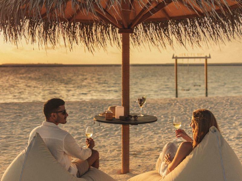 Tres personas descansan en pufs bajo una sombrilla de paja en la playa al atardecer.