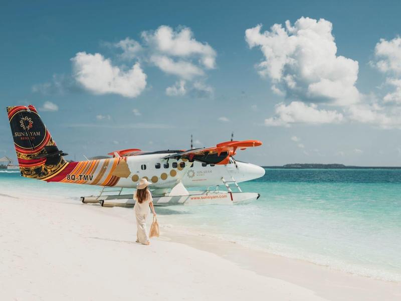 Mujer camina por la playa de arena blanca junto a un hidroavión colorido en el mar turquesa.