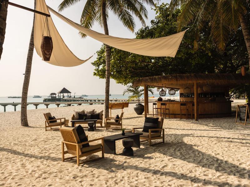 Bar de playa con sillas y mesas de madera bajo velas en entorno tropical junto al mar.