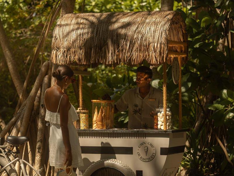 Un puesto de bebidas con techo de paja en un área sombreada con árboles y dos personas delante.