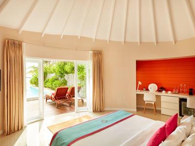 Helles Hotelzimmer mit großem Bett, Vorhängen und Blick auf eine Terrasse mit Sitzgelegenheiten.