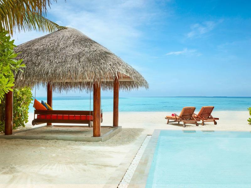 Spiaggia con palme, piscina e lettini sotto un cielo blu.