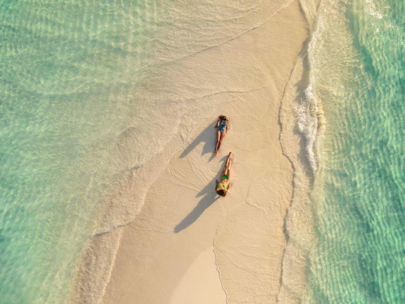 Due persone camminano su una spiaggia sabbiosa e tranquilla, circondate da acqua turchese limpida.
