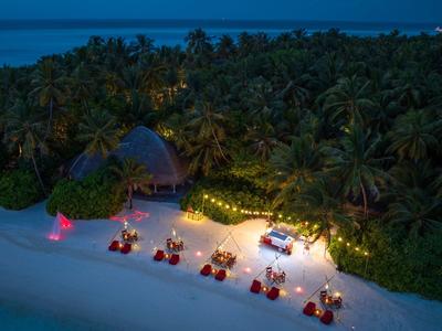 Wieczorna kolacja na plaży z oświetlonymi stolikami i leżakami przed gęstymi palmami.