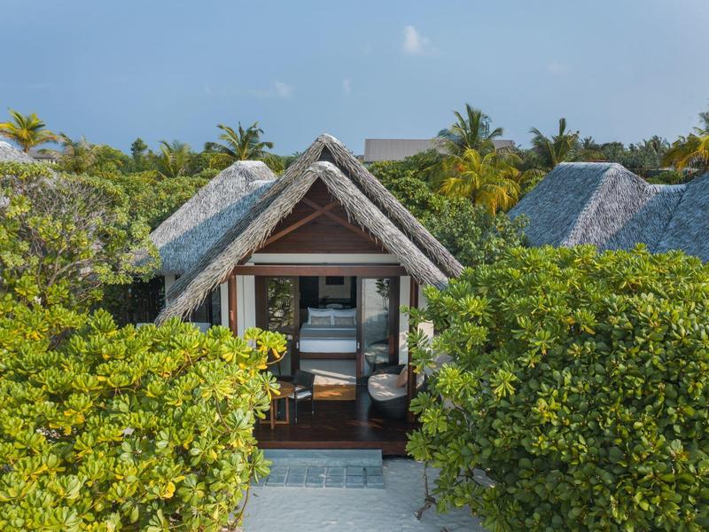 Hotelowy bungalow z dachem z trzciny otoczony gęstą zielenią i palmami pod błękitnym niebem.