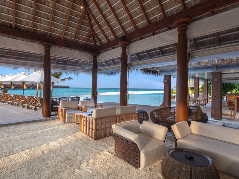 Otwarte pawilon plażowy z meblami wypoczynkowymi i widokiem na morze pod drewnianym dachem.