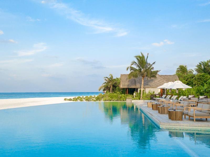 Basen infinity przy białej piaszczystej plaży z palmami i niebieską chatką pod błękitnym niebem.