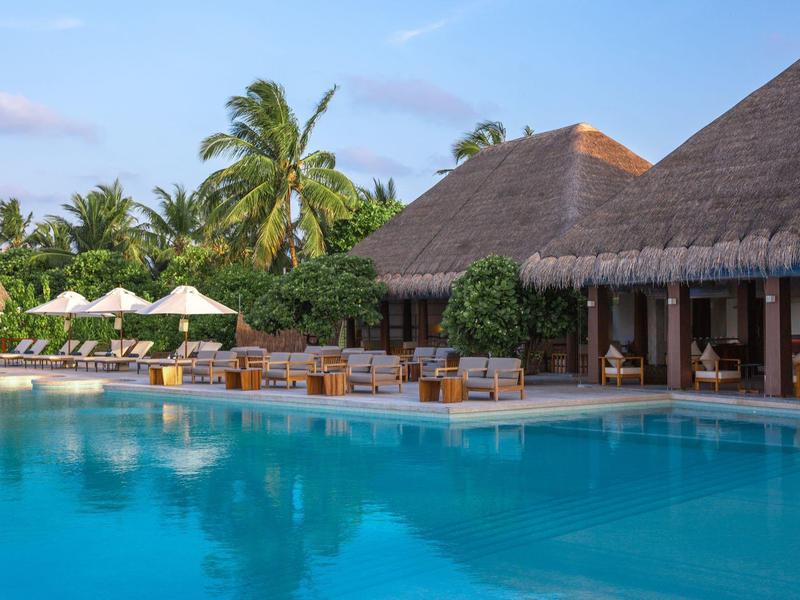 Duży basen z leżakami i parasolami przed tropikalnym budynkiem hotelu z palmami.