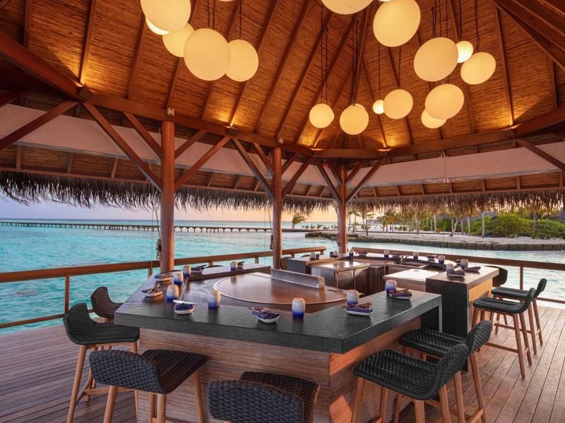 Otwarta jadalnia w ośrodku nad wodą z drewnianym stołem, krzesłami i okrągłymi wiszącymi lampami.