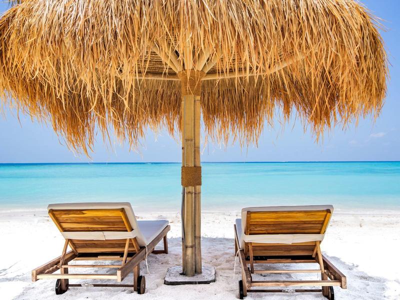 Zwei Liegestühle unter einem Strohdach am weißen Sandstrand mit Blick aufs blaue Meer.