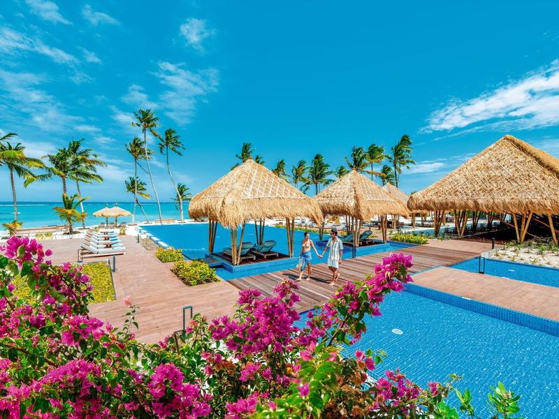 Strandresort mit Strohdächern, blauem Himmel, Palmen und bunten Blumen am Pool.