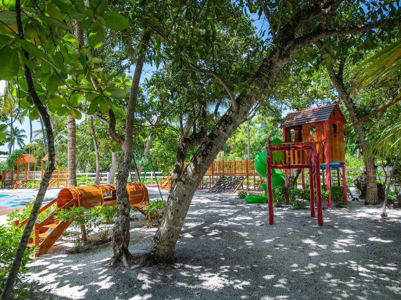 Schattenreicher Kinderspielplatz mit Klettergerüst und Rutsche unter großen Bäumen am Pool.