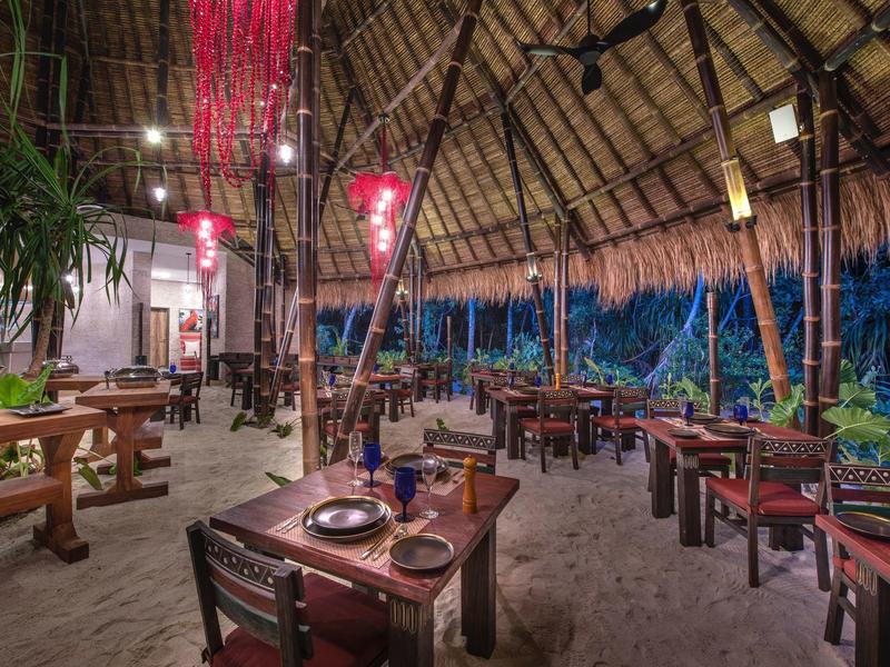 Offenes Restaurant mit Strohdach, Holzmöbeln und Sandboden in tropischem Ambiente.