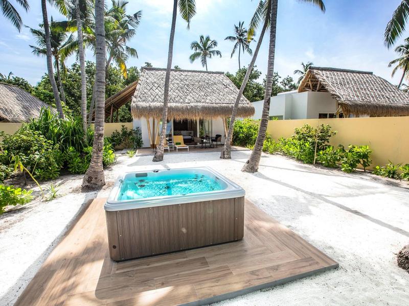 Ein Whirlpool auf einer Holzterrasse im Freien, umgeben von weißem Sand und Palmen neben einem Haus mit Strohdach.