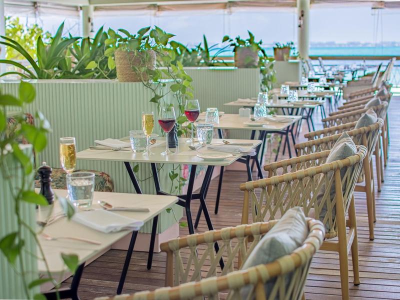 Terras met tafels en stoelen gedekt met glazen en bestek in een lichte omgeving met planten.