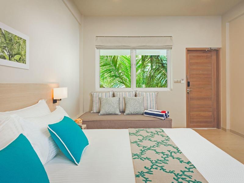 Lichte hotelkamer met tweepersoonsbed, blauw-witte kussens en uitzicht op palmbomen vanuit het raam.