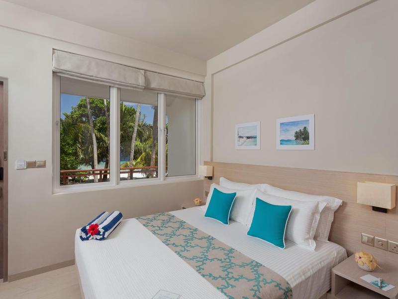 Lichte hotelkamer met tweepersoonsbed, blauwe kussens en raam met uitzicht op palmbomen.