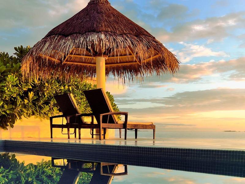 Twee ligstoelen onder een rieten parasol bij een infinity pool tijdens zonsondergang.