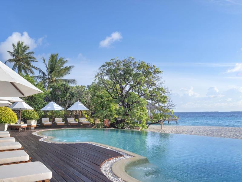 Infinityzwembad met ligstoelen, parasols en uitzicht op de oceaan onder een blauwe lucht.