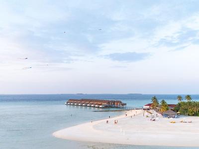 Weißer Sandstrand mit Steg ins Meer, vereinzelten Palmen und Menschen bei klarem Himmel.