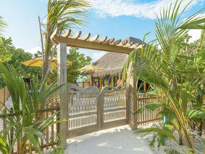 Cancello in legno con pergola in giardino tropicale, palme e cielo azzurro sullo sfondo.