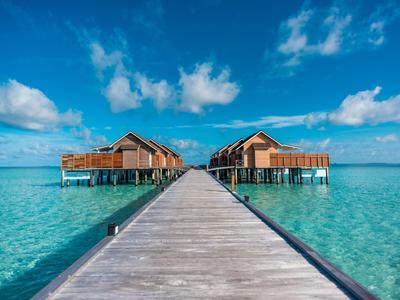 Holzsteg führt zu zwei Überwasser-Bungalows im klaren, türkisfarbenen Meer unter blauem Himmel.