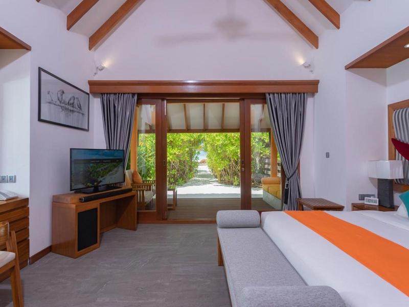 Camera d'hotel luminosa con soffitto a travi di legno, letto, TV e vista su un giardino verde.