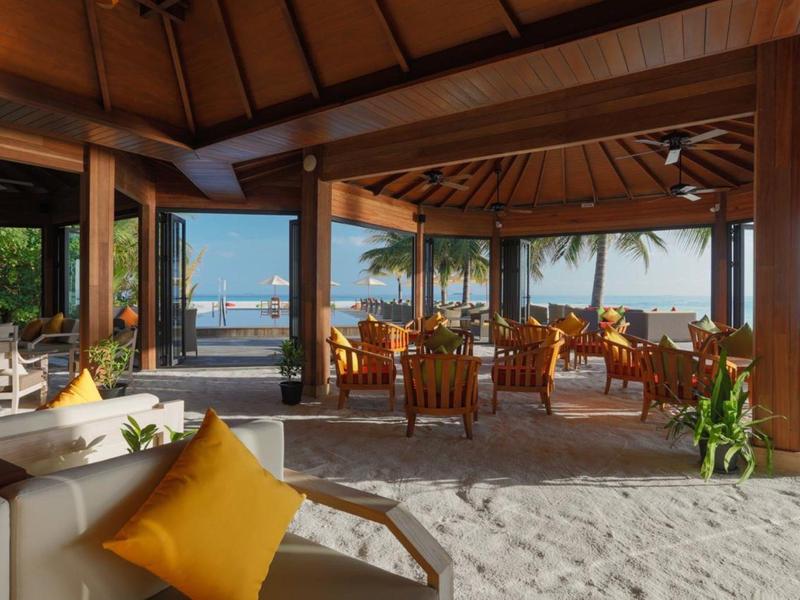 Lounge all'aperto con pavimento sabbioso, mobili in legno e vista sul mare e le palme.