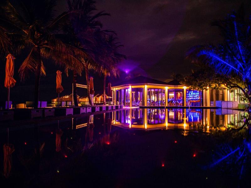 Ristorante illuminato con area esterna e palme accanto a acqua tranquilla di notte.