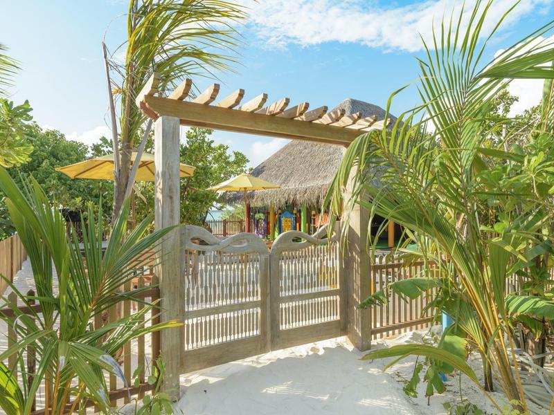 Cancello in legno con pergola in giardino tropicale, palme e cielo azzurro sullo sfondo.