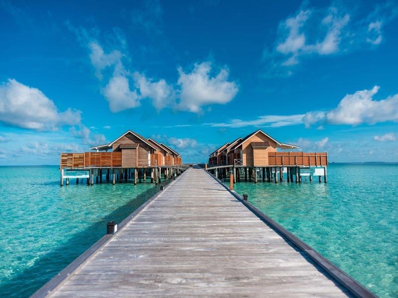 Una passerella di legno conduce a bungalow su palafitte sull'acqua cristallina sotto un cielo azzurro.