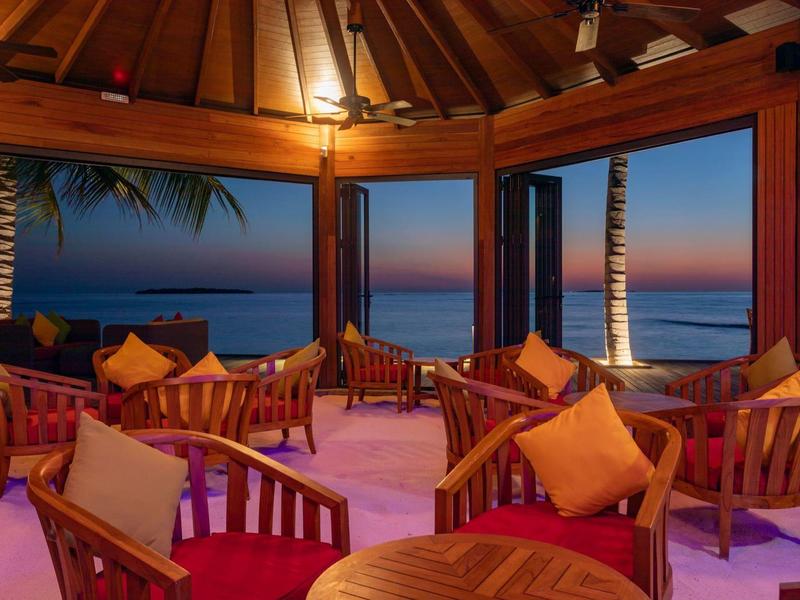 Zona lounge aperta con mobili in legno e vista palme al tramonto.