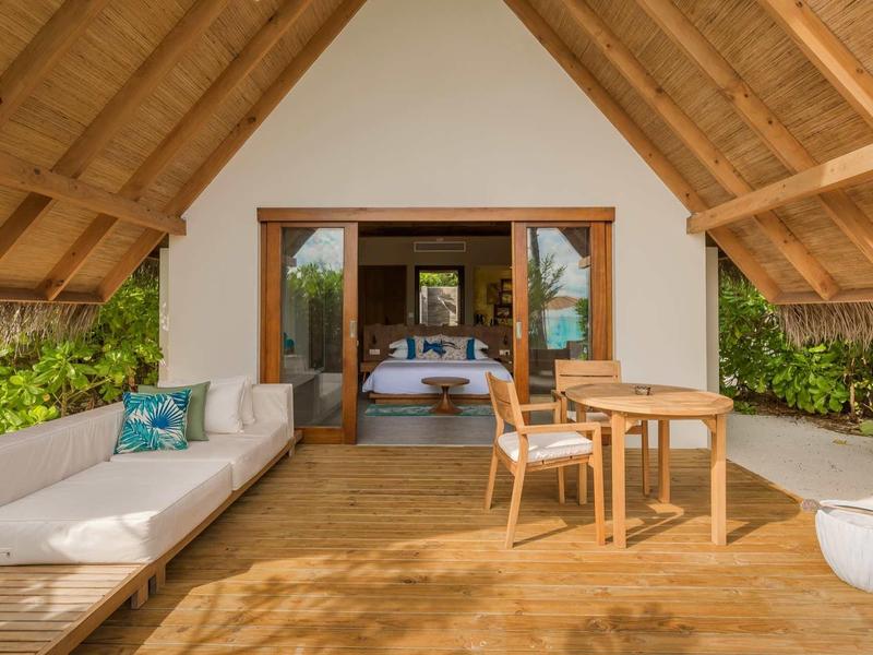 Terrassenbereich mit Holzdeck, Sofa mit Kissen, Tisch und Stühlen vor offenem Raum mit Doppelbett.