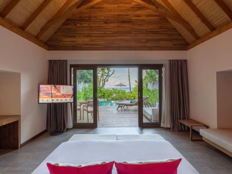 Helles Schlafzimmer mit Holzdecke, großem Bett, TV, Sofa und Blick auf Terrasse und Garten.