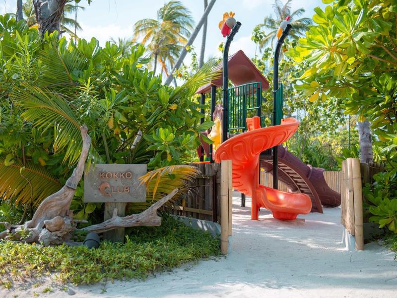 Rote Spirale-Rutsche im sonnigen, tropischen Dschungel-Spielplatz mit Palmen und Sandboden.