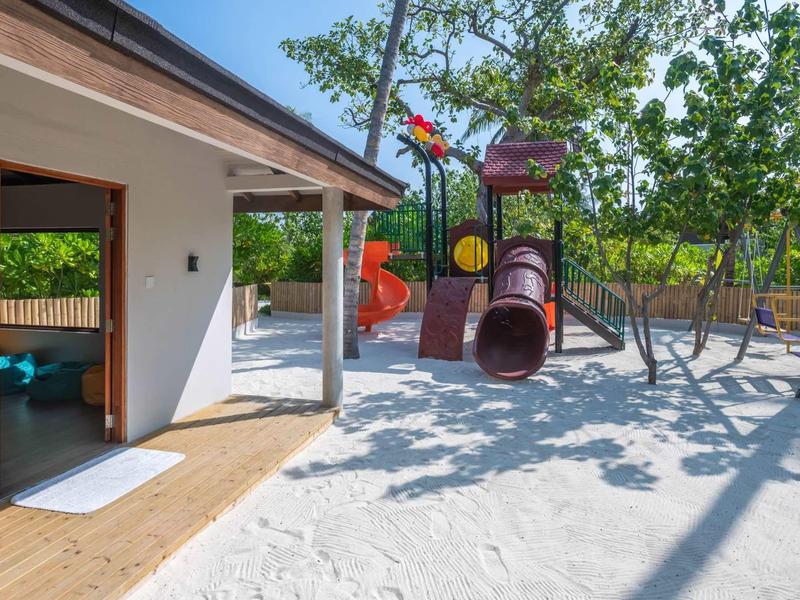 Kinderspielplatz mit Sandboden, Rutsche, Klettergerüst und angrenzendem Haus mit offener Tür.