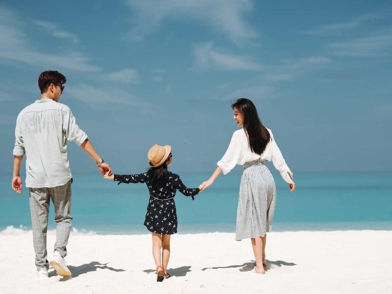 Eine Familie mit zwei Erwachsenen und einem Kind hält sich am weißen Strand an den Händen.
