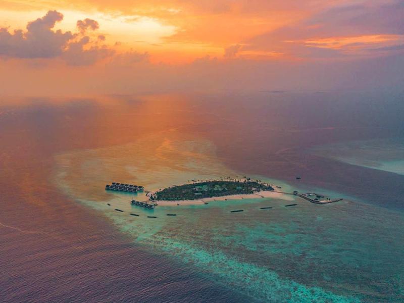 Kleine Insel mit Sandstrand und Palmen im Meer bei buntem Sonnenuntergangshimmel.